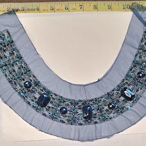 May include: A blue decorative collar with a mesh base, embellished with teal and blue rhinestones and larger blue gems. The collar is approximately 12 inches wide and is designed to be worn around the neck.