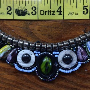 May include: A beaded collar necklace with a curved design. The necklace features a variety of beads in black, silver, orange, blue, purple, and green. The necklace is approximately 5 inches wide.