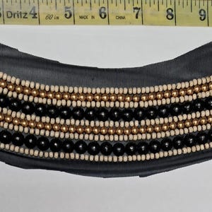May include: A black fabric collar adorned with rows of beads in black, cream, and gold. The beaded embellishments are arranged in horizontal lines across the collar. The collar is a curved shape, suitable for embellishing clothing.