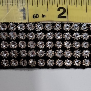 May include: Black fabric trim with rows of clear, sparkling rhinestones. The trim is measured with a yellow tape measure marked in inches. The rhinestones are set in a silver-colored metal. The trim is approximately 1 inch wide.