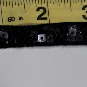 May include: Black sequin trim with a woven base, approximately 1 inch wide. The trim is shown against a white background with a yellow measuring tape indicating the width. The trim is embellished with small, square sequins.