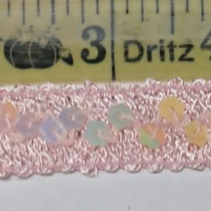May include: Pink sequin trim with a scalloped edge. The trim is approximately 1 inch wide and features iridescent sequins in shades of pink, peach, and green. The trim is measured against a yellow measuring tape.