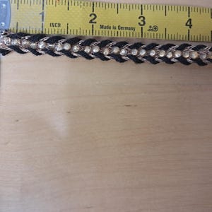 May include: Black and gold decorative trim with clear rhinestones, measuring approximately 1 inch wide. The trim is placed next to a yellow measuring tape. The trim is made in Germany.