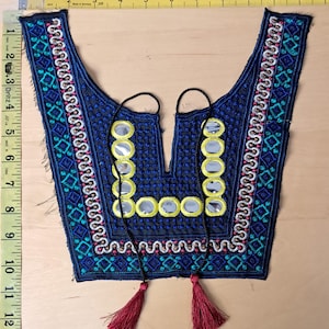 May include: A blue embroidered textile with geometric and floral patterns, featuring mirrored accents and red tassels. The textile is approximately 12 inches long, with a V-neck design and black cords.