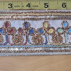 May include: Decorative trim with intricate embroidery and sequin details. The trim features a white base with gold, blue, yellow, and pink floral designs. The trim measures approximately 2 inches wide.