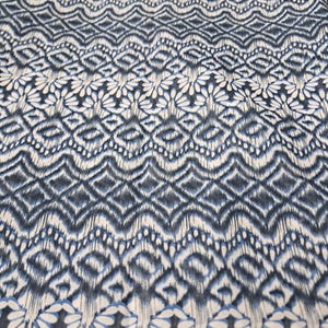 May include: A close-up of a fabric with a repeating pattern in shades of navy blue and white. The design features diamond and floral shapes, creating a textured appearance. The fabric's edge has a scalloped floral border.