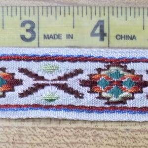 May include: A woven fabric trim featuring a repeating geometric pattern in red, teal, and dark brown on a light gray background. A yellow measuring tape is positioned along the top edge, showing the trim measures approximately 7.5 inches long.