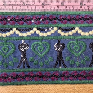 May include: Decorative trim with a denim blue background, featuring embroidered green hearts and black sequined figures. The trim is accented with rows of purple and white sequins. A pink measuring tape is in the background.