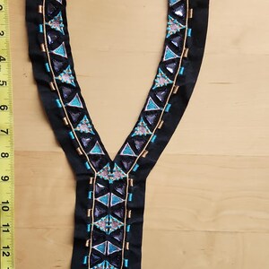 May include: Black fabric applique with a V-neck design. The applique is embellished with geometric patterns in teal, purple, and gold. The applique is approximately 14 inches long.