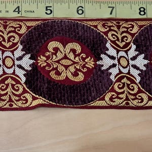 May include: Decorative trim with a repeating pattern of burgundy circles, each with a gold and red floral design. The trim is approximately 1.5 inches wide and features gold and white accents. The trim is measured with a ruler.