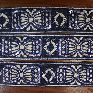 May include: Three strips of navy blue trim with white embroidery and silver sequins. The trim features a repeating pattern of a diamond shape and a bow.
