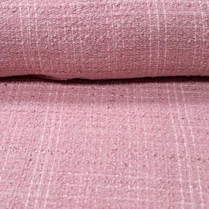 May include: Close-up of a roll of pink fabric with a textured weave. The fabric has a slightly uneven surface with a grid-like pattern of white threads. The color is a soft, muted pink, and the fabric appears to be thick and durable.