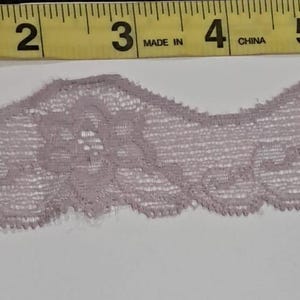 May include: A length of mauve lace with a scalloped edge, featuring a floral design. The lace is measured with a yellow tape measure, showing a width of approximately 1 inch.