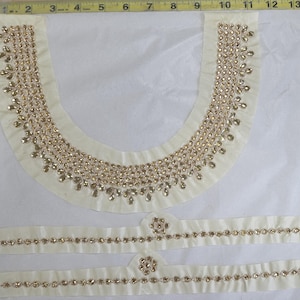 May include: An off-white fabric neckline applique and two matching sleeve bands embellished with gold-toned teardrop and round crystals. A yellow measuring tape shows the neckline is approximately 10 inches wide and 8 inches deep.