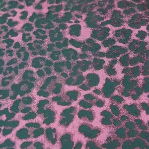 May include: Close-up of a pink fabric with a dark green leopard print pattern. The pattern consists of irregular, dark green spots scattered across the pink background, creating a textured appearance. The fabric has a subtle sheen.
