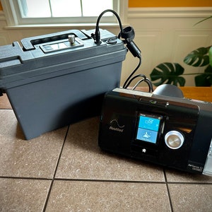 May include: A black and gray portable oxygen concentrator with a digital display and a clear water tank. The concentrator is connected to a black battery pack in a gray plastic case.