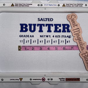 May include: An embroidery hoop with the words "SALTED BUTTER" in blue. A pink measuring tape shows approximately 7 inches. A wooden sign says "Sweet Magnolia Blooms". The net weight is 4 oz (113.4g).