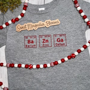 May include: Gray t-shirt with the text "Sweet Magnolia Blooms" and three periodic table elements: Barium (Ba), Zinc (Zn), and Gallium (Ga).