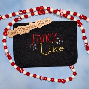 May include: A black fabric pouch with the text "Sweet Magnolia Blooms" on a wooden tag and the text "FANCY Like" embroidered in red and gold thread. The pouch is surrounded by a red and white beaded garland.