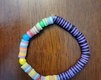May include: A colorful bracelet made of stacked, striped, and solid round beads. The bracelet is half black and purple and half rainbow colors.