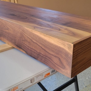 May include: A long, rectangular floating shelf made of dark walnut wood. The shelf is resting on a white folding table. The wood grain is visible, and the edges are cleanly finished. The shelf is approximately 72 inches long.