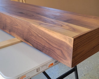 May include: A long, rectangular floating shelf made of dark walnut wood. The shelf is resting on a white folding table. The wood grain is visible, and the edges are cleanly finished. The shelf is approximately 72 inches long.