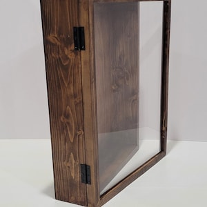 May include: A dark brown wooden display case with a glass front and black metal hinges. The case is closed and standing upright on a white surface.