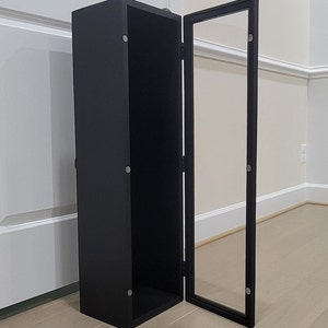 May include: A black cabinet with a glass door. The cabinet has a black interior and is closed. The door is open and the cabinet is standing on a light wood floor.
