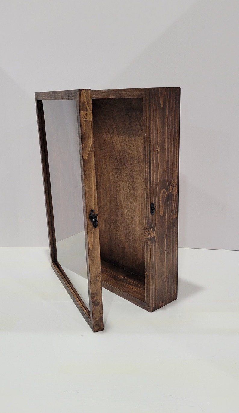 May include: A dark brown wooden display case with a glass front and a hinged door. The case is open and the door is propped open at a 45-degree angle. The case has a simple design with a smooth finish.