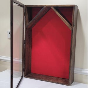 May include: A wooden display case with a hinged glass door, open to the side. The interior is lined with red fabric. The frame is made of dark stained wood.
