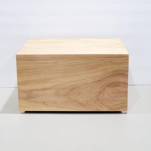 May include: A natural wood box with a light finish. The box is a cube shape and sits on four small, white feet. The wood grain is visible on the sides and top of the box.