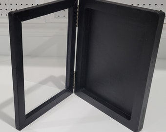 May include: Black wooden picture frame with a hinged design. The frame is open and the back is visible.