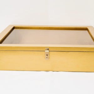 May include: A gold wooden display box with a glass top and a metal latch. The box is closed and the latch is locked.