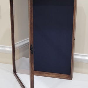 May include: A wooden display case with a hinged glass door, open to reveal a dark blue interior. The frame is a rich brown color, and the case is designed to showcase collectibles or memorabilia. The case is sitting on a white surface.
