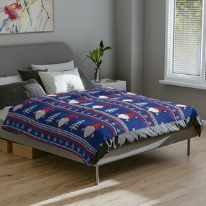 May include: A blue blanket featuring a repeating pattern of white gnomes with red hats, snowflakes, and Christmas trees. The blanket is on a bed with a grey headboard and wooden floor.