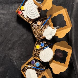 May include: Four brown cardboard boxes with clear plastic windows, each containing a white ceramic craft item and paint pots. The items are a menorah, a dreidel, a Star of David, and a small angel.