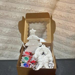 May include: A brown cardboard box with a clear window containing two white ceramic ornaments, one shaped like a Christmas tree and one like a candy cane with a bow, nestled in white paper shreds with small containers of red, green, and blue paint.