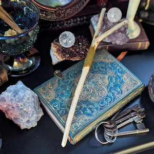 May include: An assortment of mystical items, including a blue and gold embossed journal, crystals, a wand, and keys. A clear glass bowl with cinnamon sticks sits near a collection of antique books. The items are arranged on a dark surface.