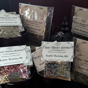 May include: Various candle dressing kits in clear bags, each with a label. The kits contain dried herbs and botanicals. Labels include text such as "Samhain," "Ostara," and "Yule." The kits are displayed in small, dark-coloured bowls.