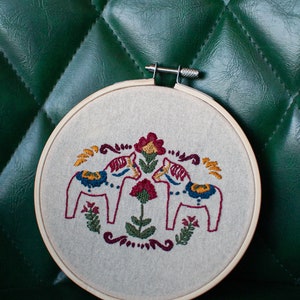 May include: Embroidered hoop art featuring two white Dala horses facing each other with red, yellow, blue, and green details. The horses are surrounded by a floral design with red, green, and yellow flowers.