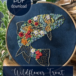 Puede incluir: Arte de bordado en aro con una trucha con un diseño floral. La trucha está delineada con hilo dorado y llena de flores coloridas. El fondo es una tela azul oscuro. El texto "Wildflower Trout by Wyo Wildflower" está en la parte inferior del aro.