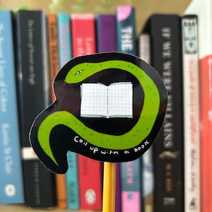 Puede incluir: Una pegatina en un lápiz con una serpiente verde enroscada alrededor de un libro abierto. La pegatina tiene el texto "Coil up with a book" en blanco. Libros de varios colores en el fondo.