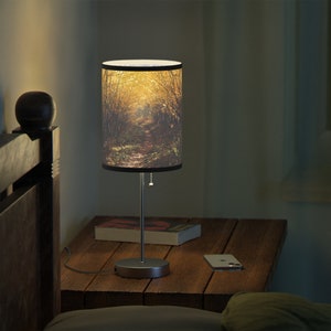 May include: A table lamp with a cylindrical lampshade featuring a photograph of a path through a forest. The lampshade is brown and has a black trim. The lamp base is silver and has a round base.