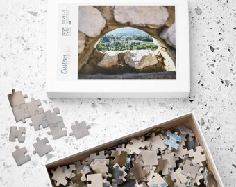 Può includere: Una scatola bianca con un'immagine di un arco in pietra con vista su una valle verde attraverso l'arco. La scatola è etichettata "Custom Made" e "Devel 2". La scatola è appoggiata su una superficie bianca con un mucchio di pezzi di puzzle sparsi intorno.