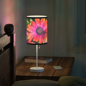 May include: A white table lamp with a black and white lampshade featuring a pink and orange daisy with water droplets. The lamp is on a wooden table with a book and a phone.