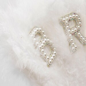 May include: Two white letters, "B" and "R", made of pearls and rhinestones, on a white fluffy background.