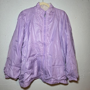 May include: A light purple jacket with a full zipper closure, two front pockets, and a stand-up collar. The jacket is hanging on a wooden hanger against a white wall. The jacket appears to be made of a shiny, lightweight material.