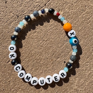 May include: A circular beaded bracelet featuring white letter beads spelling out OK COMPUTER, interspersed with various round beads in shades of blue, grey, black, orange, and clear, including a blue and white evil eye bead and a white bead with an X.