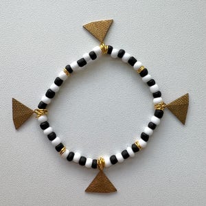 May include: A beaded bracelet featuring alternating matte black and white spherical beads arranged in a circular pattern. The design is accented with four gold-toned triangular charms with a textured surface, each attached to the band with gold wire.