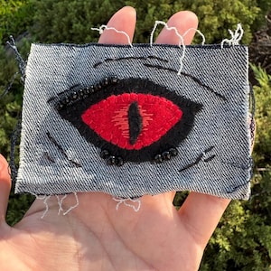 May include: A rectangular denim patch featuring an embroidered eye design. The eye has a bright red iris with a vertical black pupil, surrounded by black stitching and small black beads. The denim fabric has frayed edges and black thread accents.